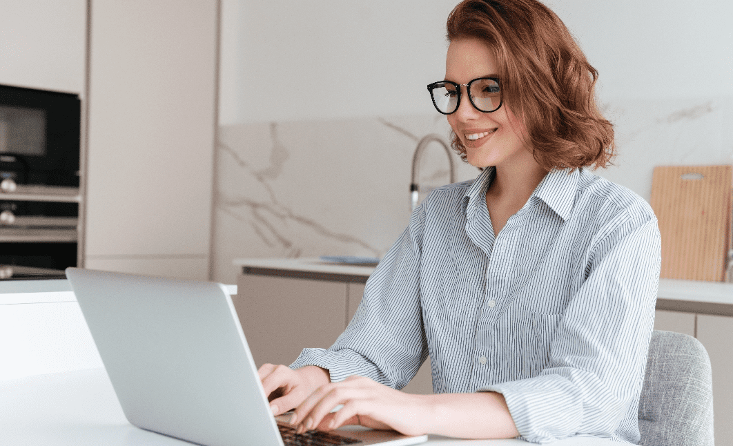 elegante-mujer-sonriente-gafas-camisa-rayas-usando-computadora-portatil-mientras-sienta-mesa-cocina@2x